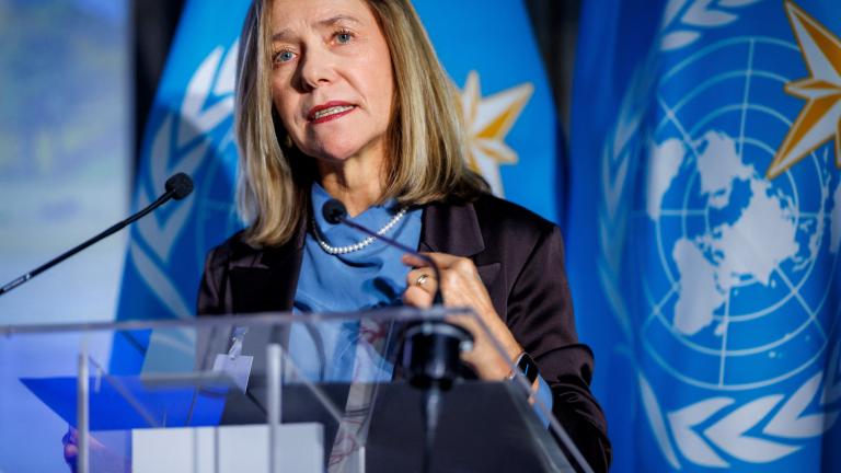 A woman speaks at a podium with microphones, standing in front of blue flags featuring a world map and laurel wreath symbol.
