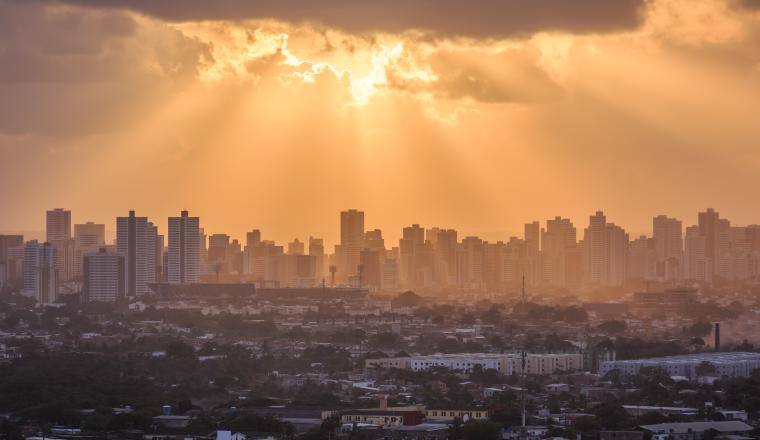 Sun rays break through clouds over a city skyline with high-rise buildings, casting a warm golden light on the urban landscape below.