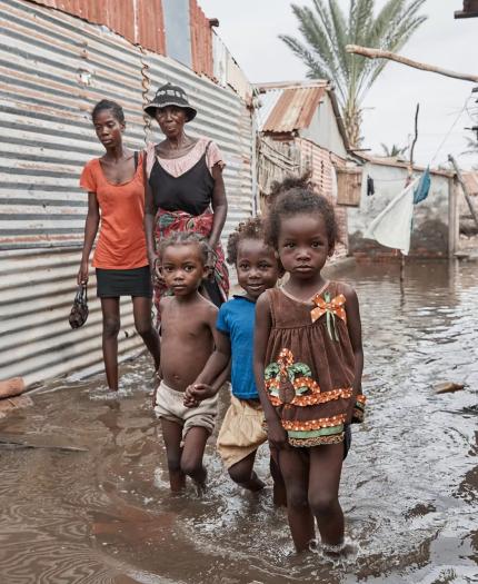 Children and adults stand in a flooded alleyway between makeshift homes made of corrugated metal and natural materials.