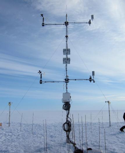Two scientists working on a complex weather station apparatus on a snow-covered plain under a clear blue sky.