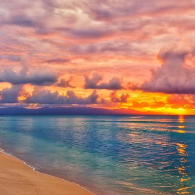 A sandy beach with calm blue water at sunset, palm trees on the left, and colorful clouds in the sky reflecting orange and pink hues.