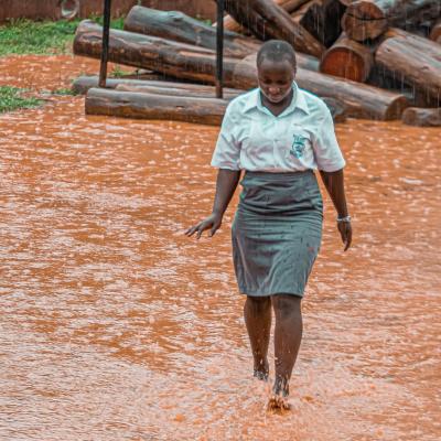 A student in uniform walks barefoot through a flooded, muddy area during heavy rain, with stacked logs in the background.