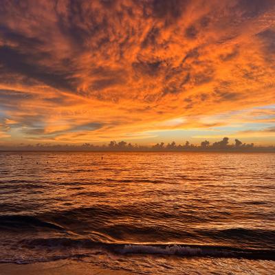 Orange and gold clouds fill the sky over a calm ocean at sunset, reflecting warm colors on the water and gentle waves near the shore.