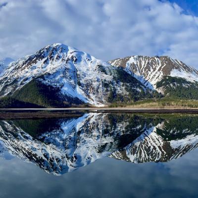 Snow-capped mountains are reflected in a calm lake under a blue sky with scattered clouds.