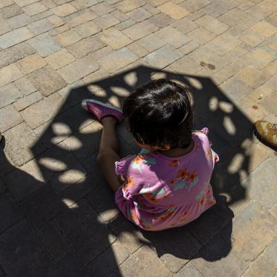 A child in a pink floral dress sits on a stone pavement, partially shaded by an umbrella, with three adults’ legs visible nearby.
