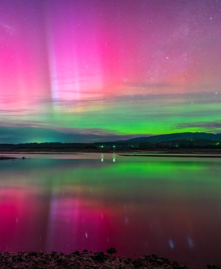 Vibrant aurora borealis reflected over a tranquil lake, with pink and green hues in the sky and a serene landscape in the background.