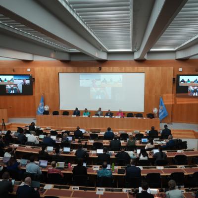 A conference room with attendees seated at desks facing a panel of speakers and a large screen, with United Nations flags displayed on both sides of the stage.