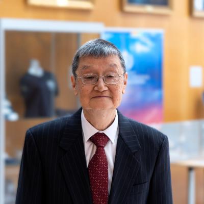 An elderly man in a dark pinstripe suit, white shirt, and red tie stands indoors with display cases and framed pictures in the background.