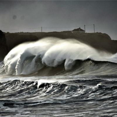 Large ocean waves crash near a rocky coast under a cloudy sky, with a silhouette of a cliff and buildings in the background.