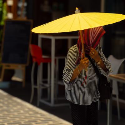 Person holding a large yellow umbrella, face covered by a striped scarf, standing on a sunlit sidewalk near outdoor seating and a blackboard sign.