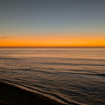 Calm ocean waves under a clear sky at sunset, with an orange glow on the horizon and silhouettes of land on the left side.