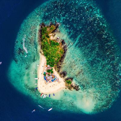 Aerial view of a small, lush island surrounded by a shallow coral reef and anchored boats.